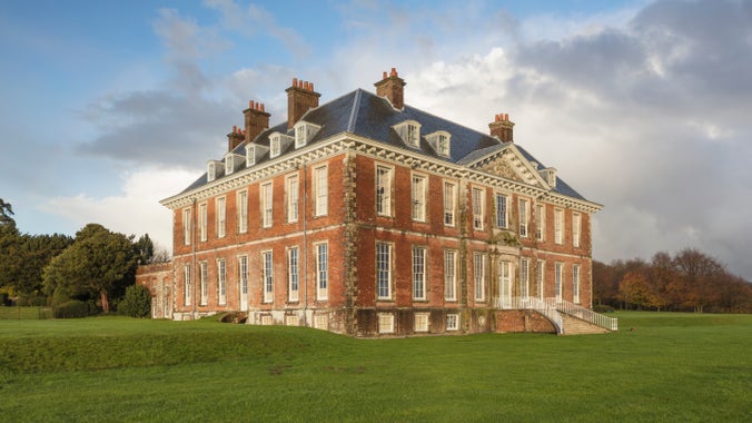 A corner view of the exterior of Uppark House, West Sussex, in red brick with many windows, standing in grassland